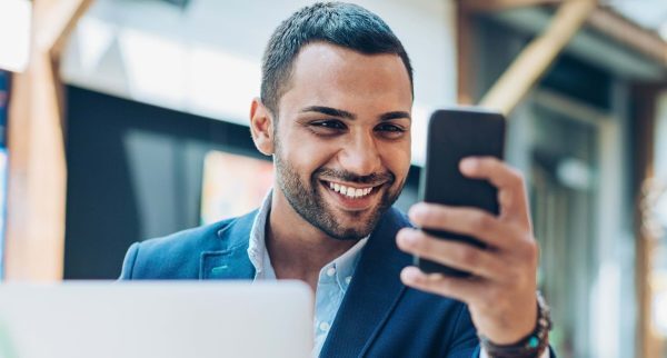 Smiling young man sitting in restaurant and texting, with his laptop in front of him.